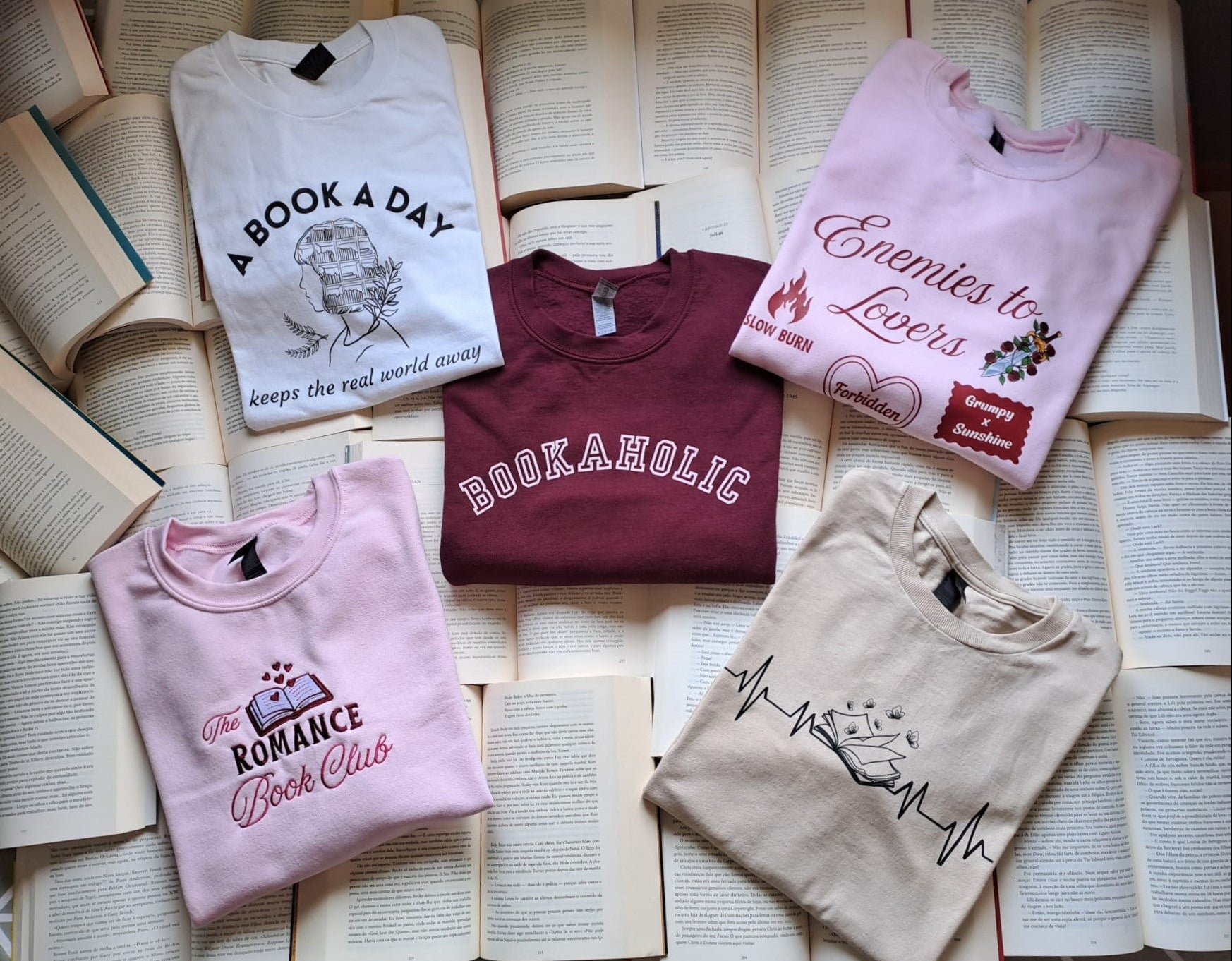 Five sweatshirts with book-themed designs on a surface of open books
